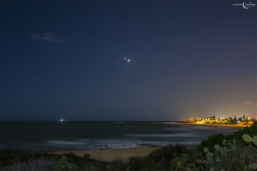 Two bright objects are pictured very near each other in night sky
filled with stars. A beach is in the foreground, with some lit structures
visible across the water. 
Please see the explanation for more detailed information.