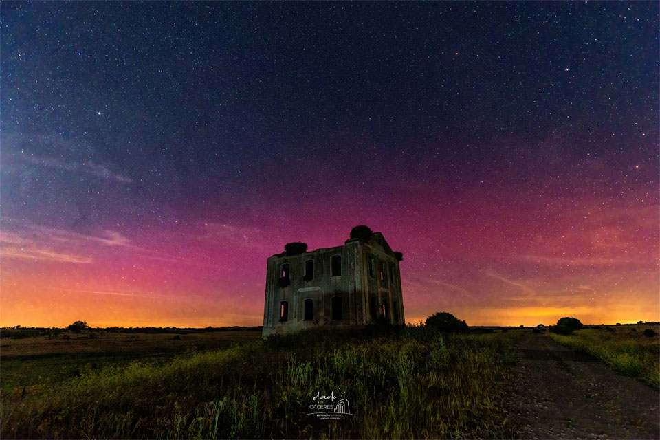 A dark rectangular building is seen across a green field
with colorful aurora, mostly red, seen in the background sky. 
Past the aurora, the sky is also filled with stars. 
Please see the explanation for more detailed information.