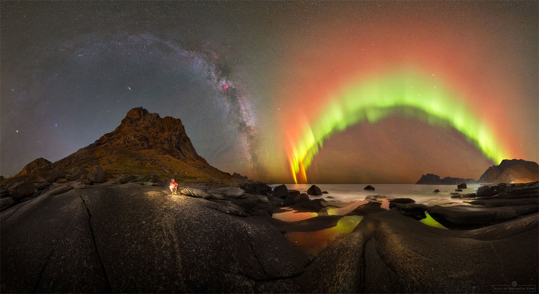 A night sky filled with stars is shown behind a picturesque foreground.
The foreground contains rounded rocks and a person before a distant sea.
The background contains bands of the Milky Way and bright aurora.
Please see the explanation for more detailed information.