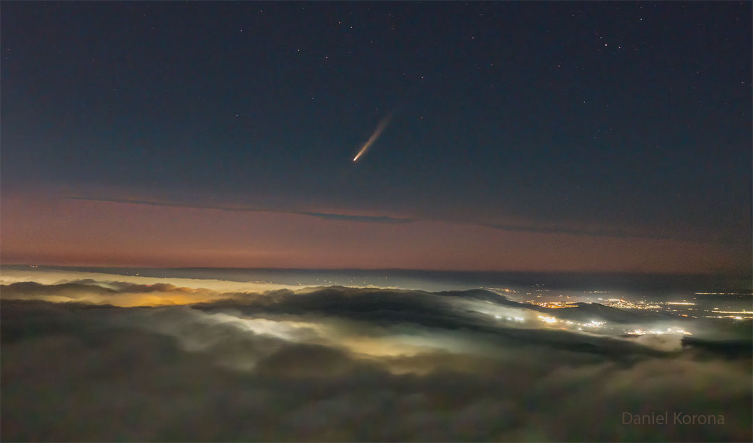 A picture shows clouds across the bottom and a dark night sky
across the top. In the middle is a band of orange sky. City lights
are visible on the right through gaps in the clouds. In the center
of the upper sky is a comet with its tail pointing toward the 
upper right. 
Please see the explanation for more detailed information.