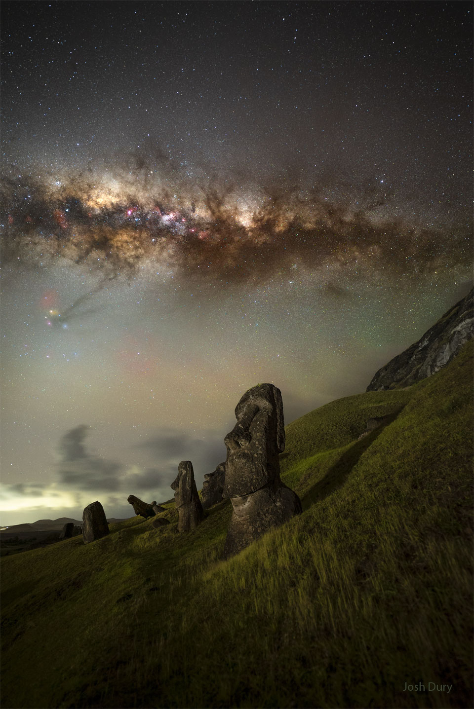 A grassy hill appears in the foreground with tall statues 
of human heads embeddd. High overhead the central band of the 
Milky Way galaxy crosses horizontally. Above the Milky Way is
a dark sky filled with stars. 
Please see the explanation for more detailed information.