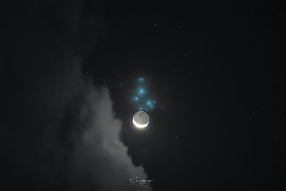A dark night sky is shown with clouds on the left. 
Just above center in blue is the Pleiades star cluster. 
Just below the Pleiades is a crescent moon, but bright 
enough so that you can see not only the brightly lit
crescent but, more faintly, the rest of the Moon.
Please see the explanation for more detailed information.